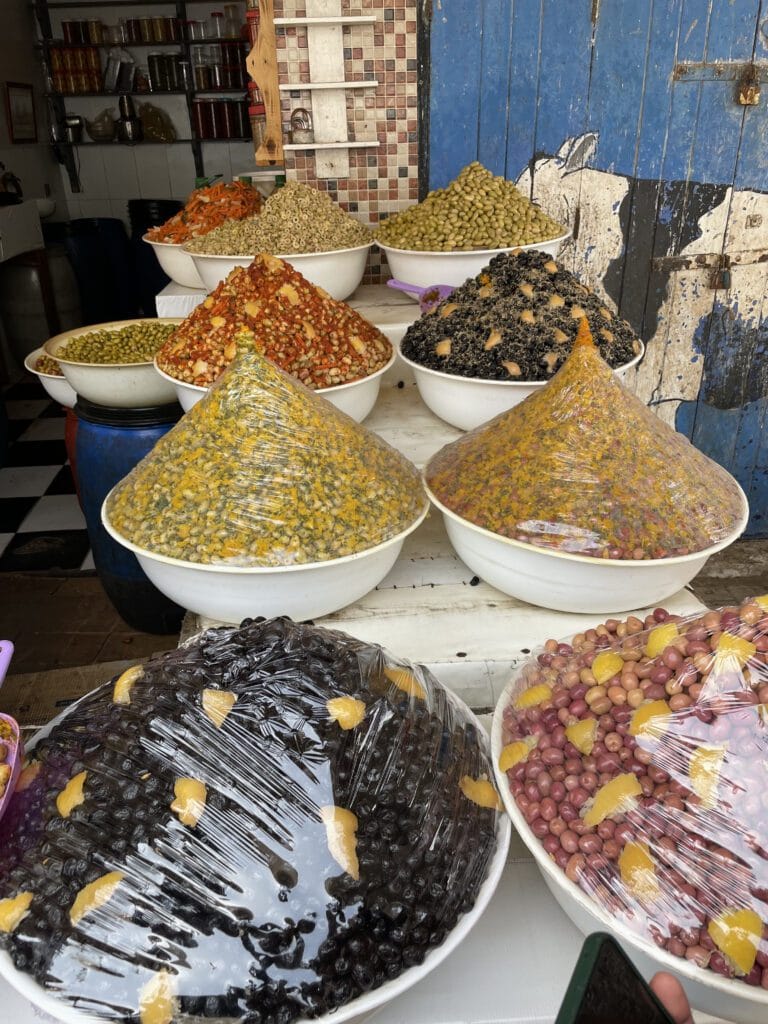 A selection of diverse foods in bowls, including olives, displayed at a vibrant market setting.