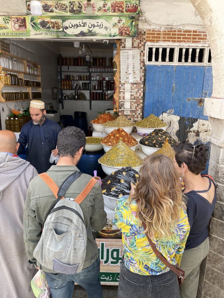 A group of people gathered around a food stand at a market, examining various olive products on display.